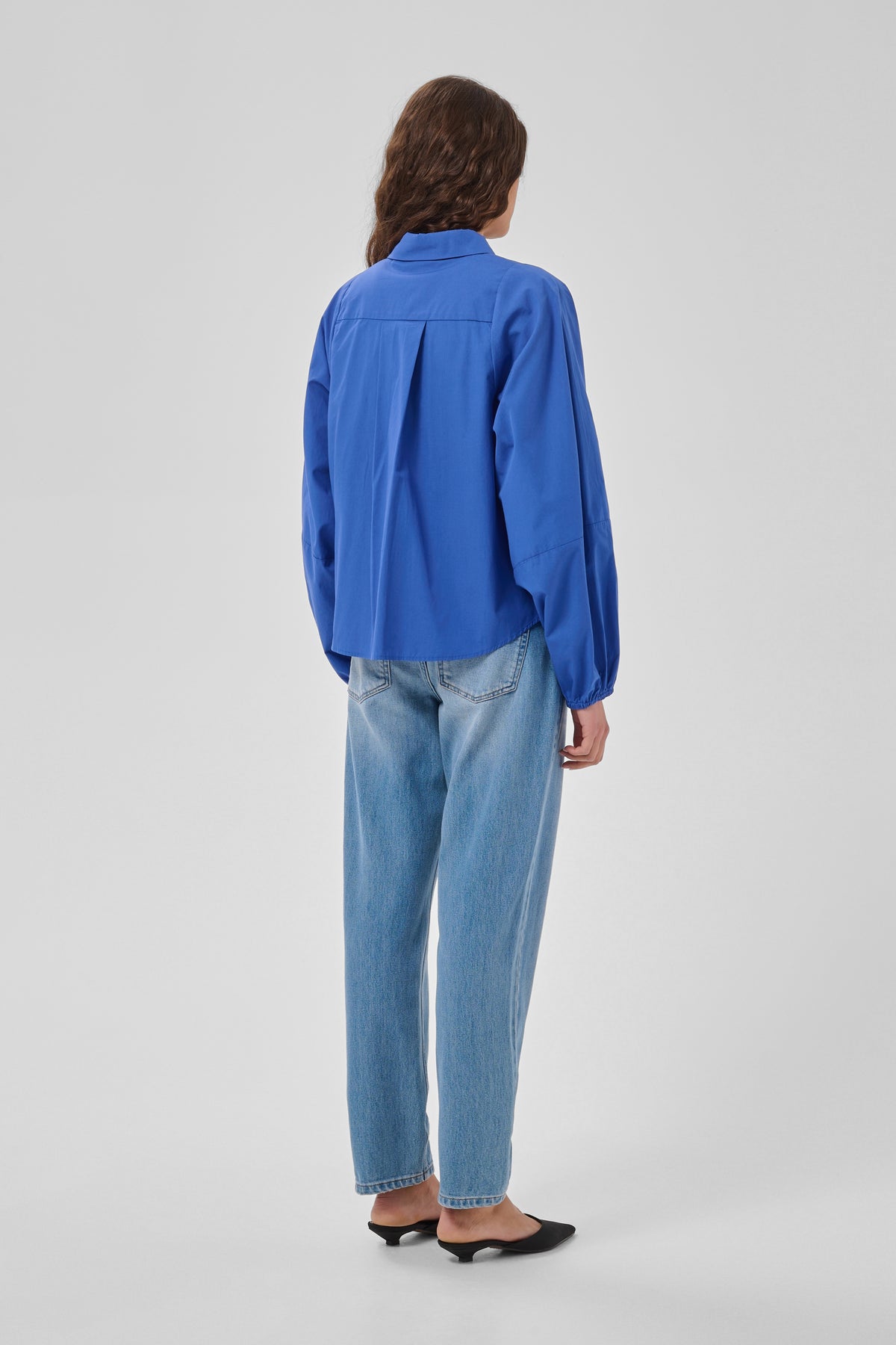 A model shown from the back wearing a bright blue long-sleeve button-up shirt and light-wash blue jeans, paired with black pointed flats, against a light gray studio background.