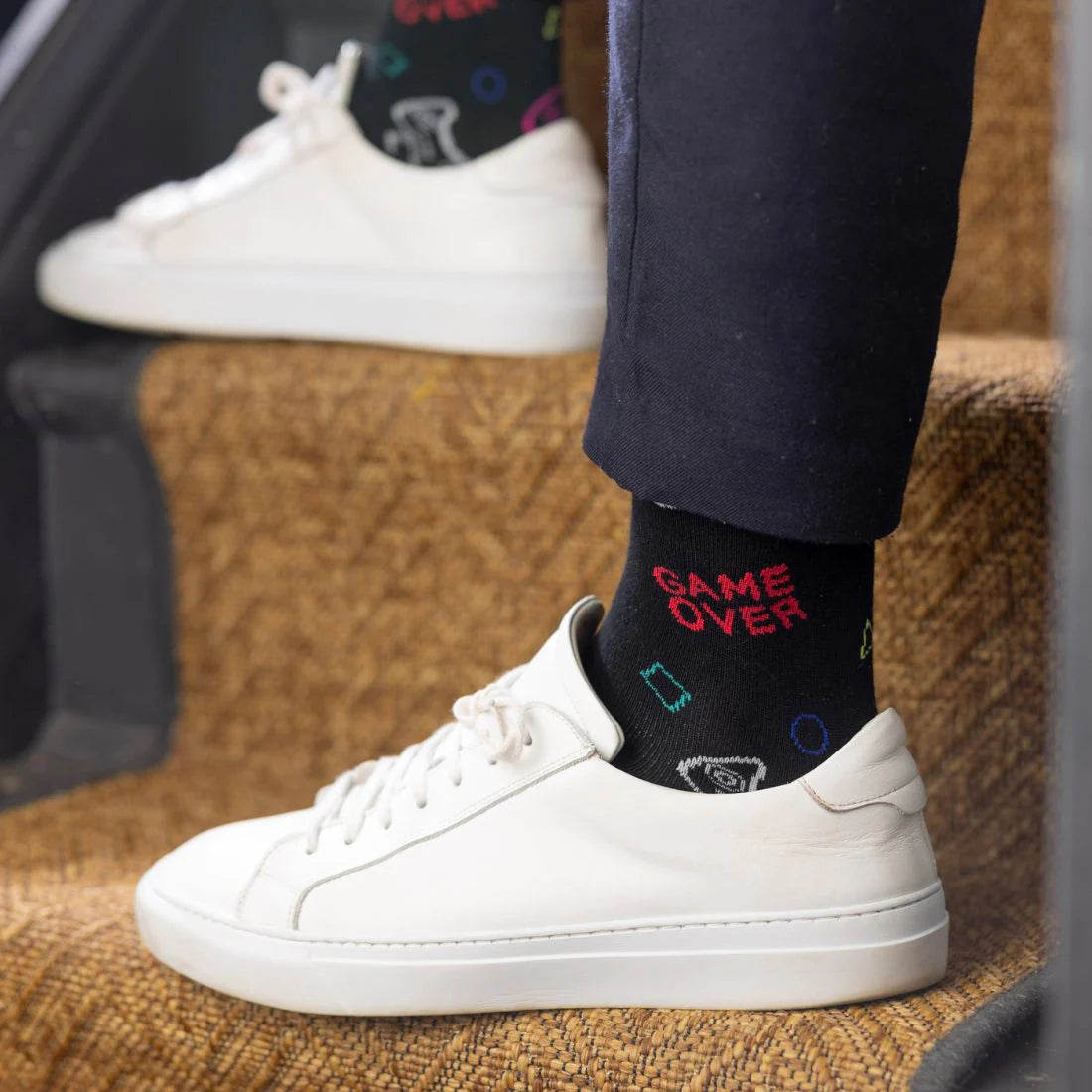 Person wearing black socks with 'GAME OVER' text and white sneakers on a textured surface.