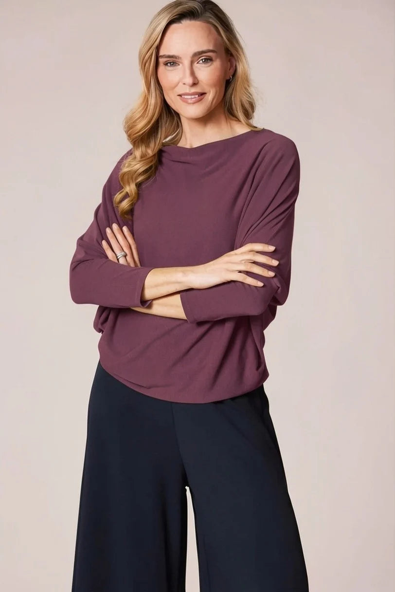 A woman modeling a long-sleeve boat-neck top in a muted plum purple color, paired with dark navy wide-leg pants, against a solid light beige studio background.