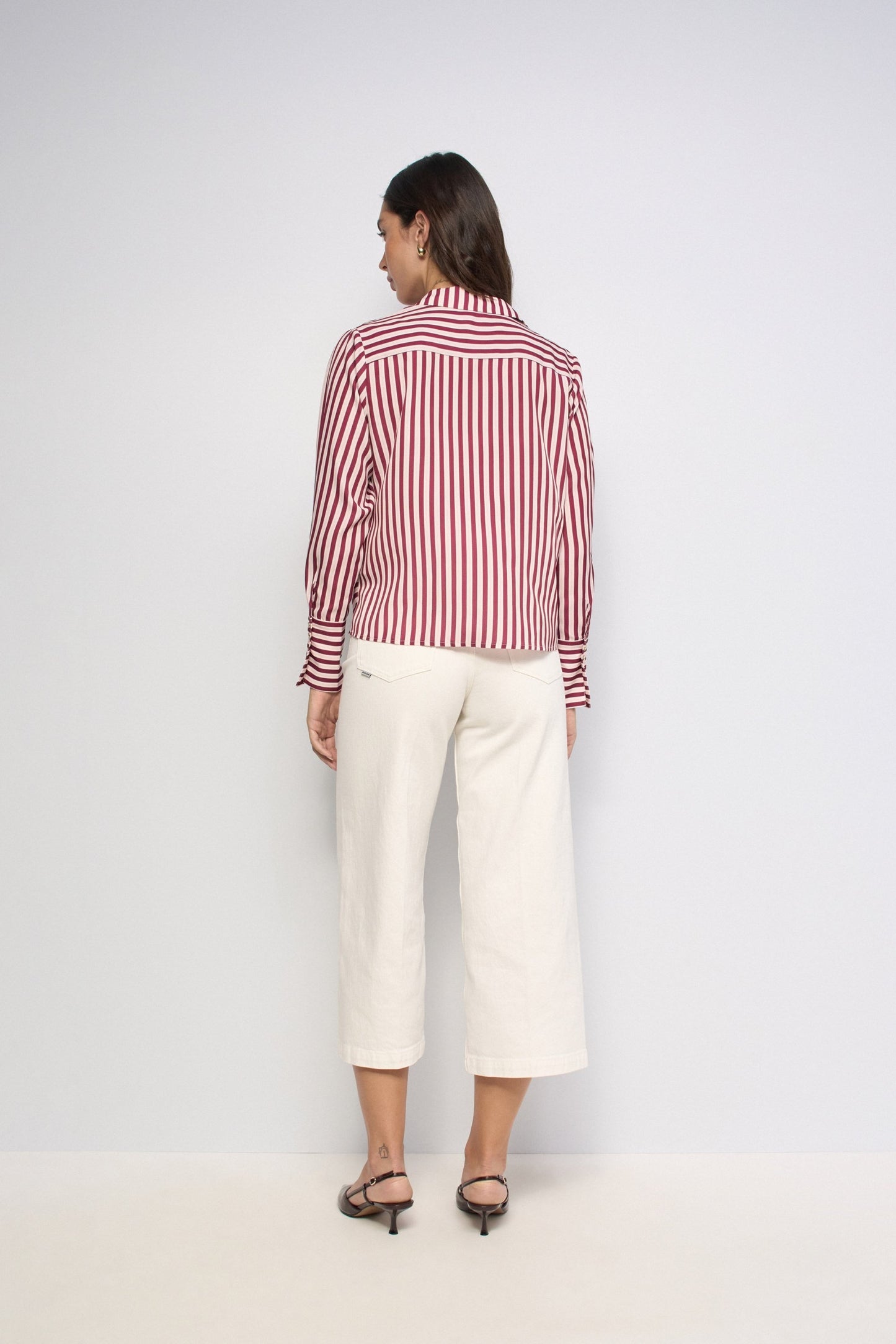 A woman is shown from the back wearing a red and white vertically striped long-sleeve button-up shirt and off-white cropped trousers, paired with black heeled sandals, against a plain light gray studio background.
