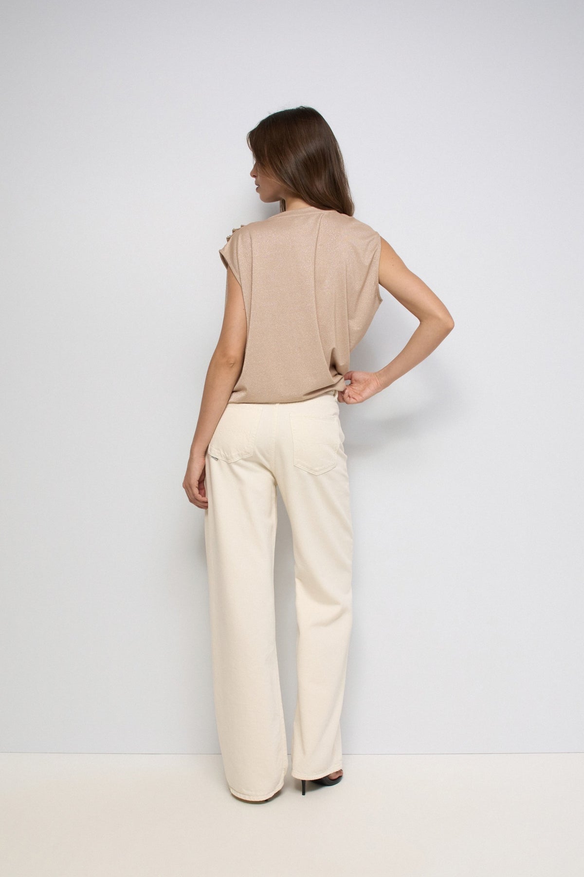 A woman shown from the back wearing a beige sleeveless top and cream wide-leg trousers, standing against a plain light gray studio background.