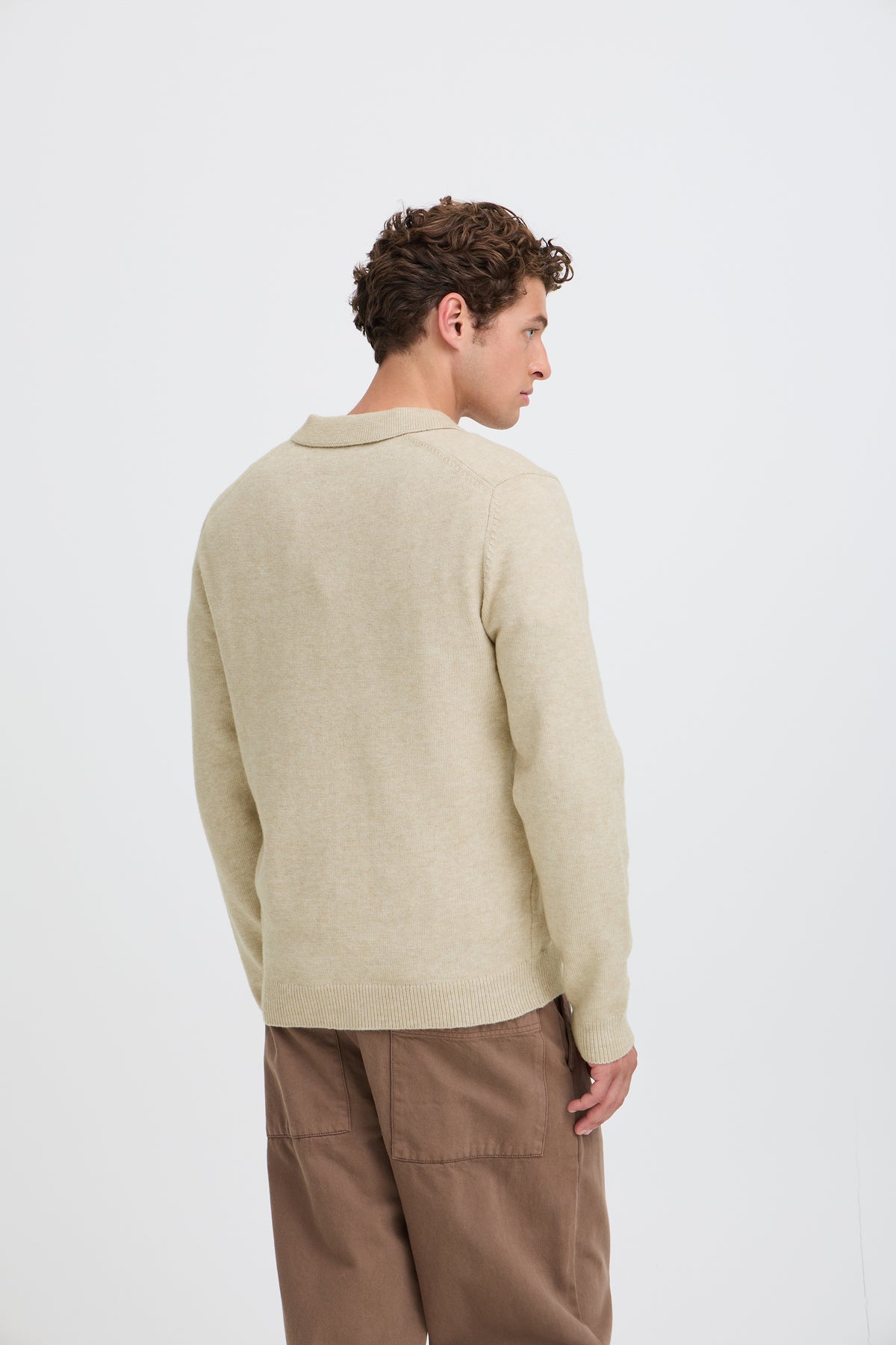 A man wearing a light beige knit polo sweater and brown trousers, photographed from the back against a clean white studio background.