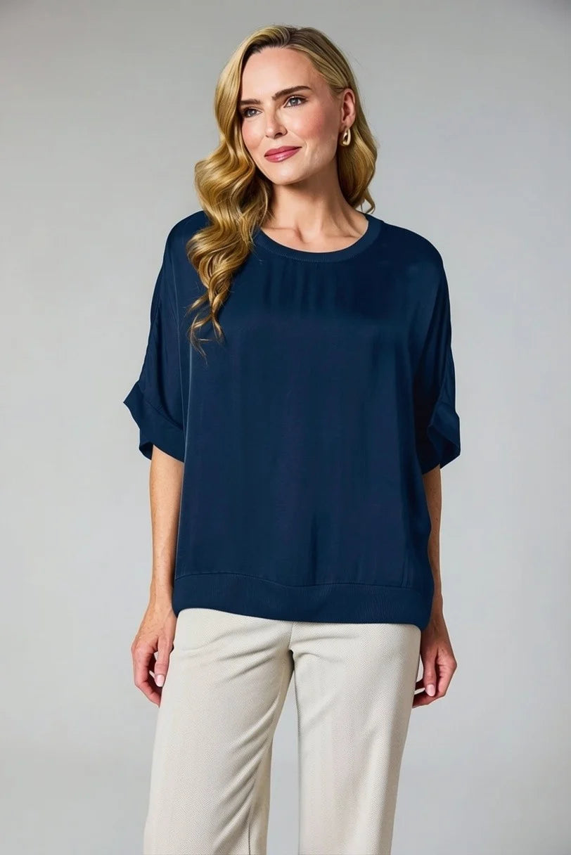 A woman modeling a navy blue short-sleeve top with rolled cuffs and light beige pants, standing against a neutral gray studio background.