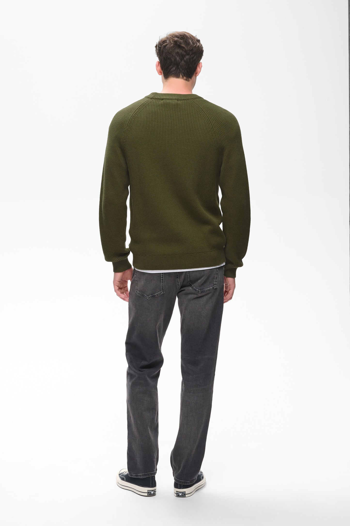 A man viewed from behind wearing an olive green crewneck sweater, dark gray jeans, and black sneakers, standing against a plain white studio background.