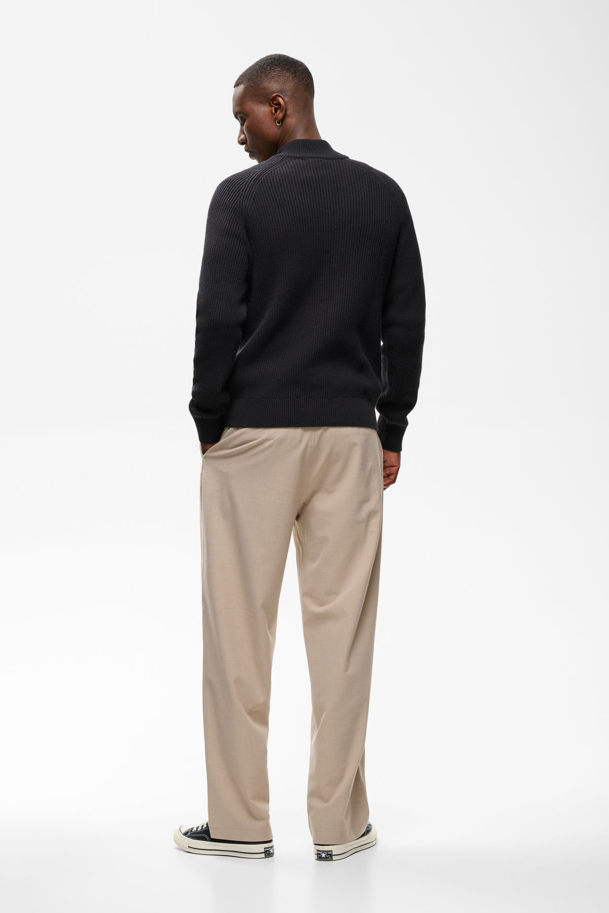 A man viewed from behind wearing a black long-sleeve knit sweater, beige trousers, and black-and-white sneakers, standing against a clean white studio background.