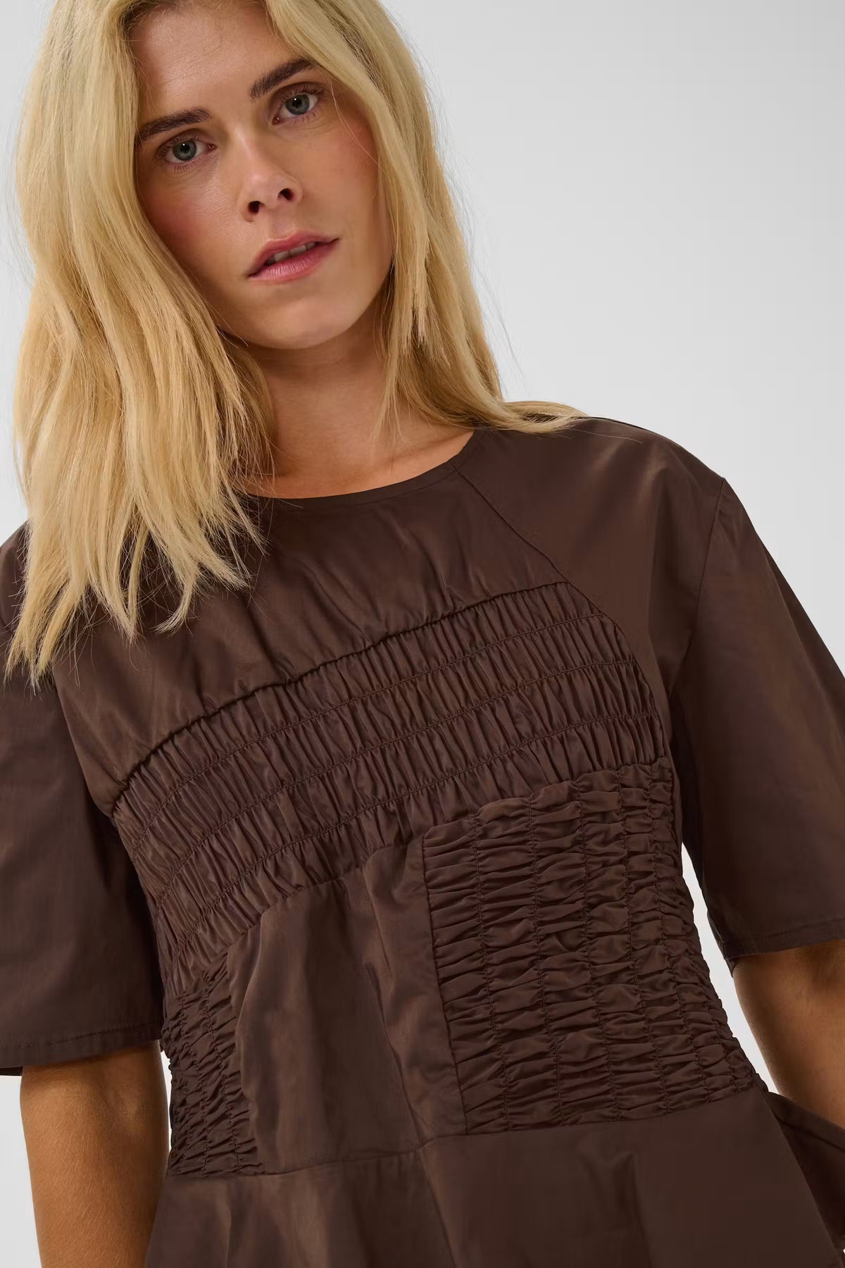 A close-up of a woman wearing a brown short-sleeve top with a smocked bodice and gathered skirt details, photographed against a light gray studio background.