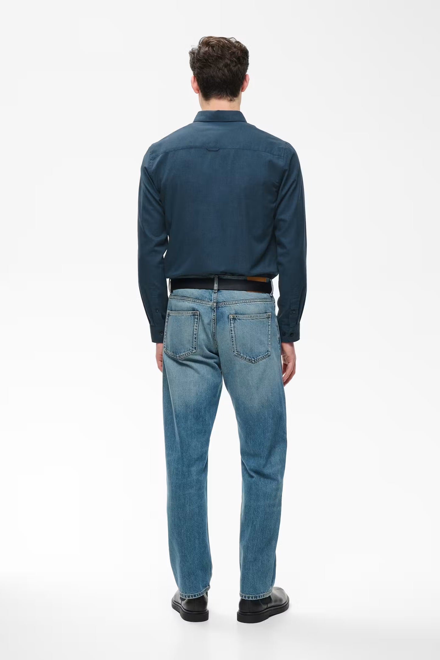 A man viewed from behind wearing a dark blue long-sleeve button-up shirt, blue jeans, a black belt, and black shoes, standing against a plain white studio background.