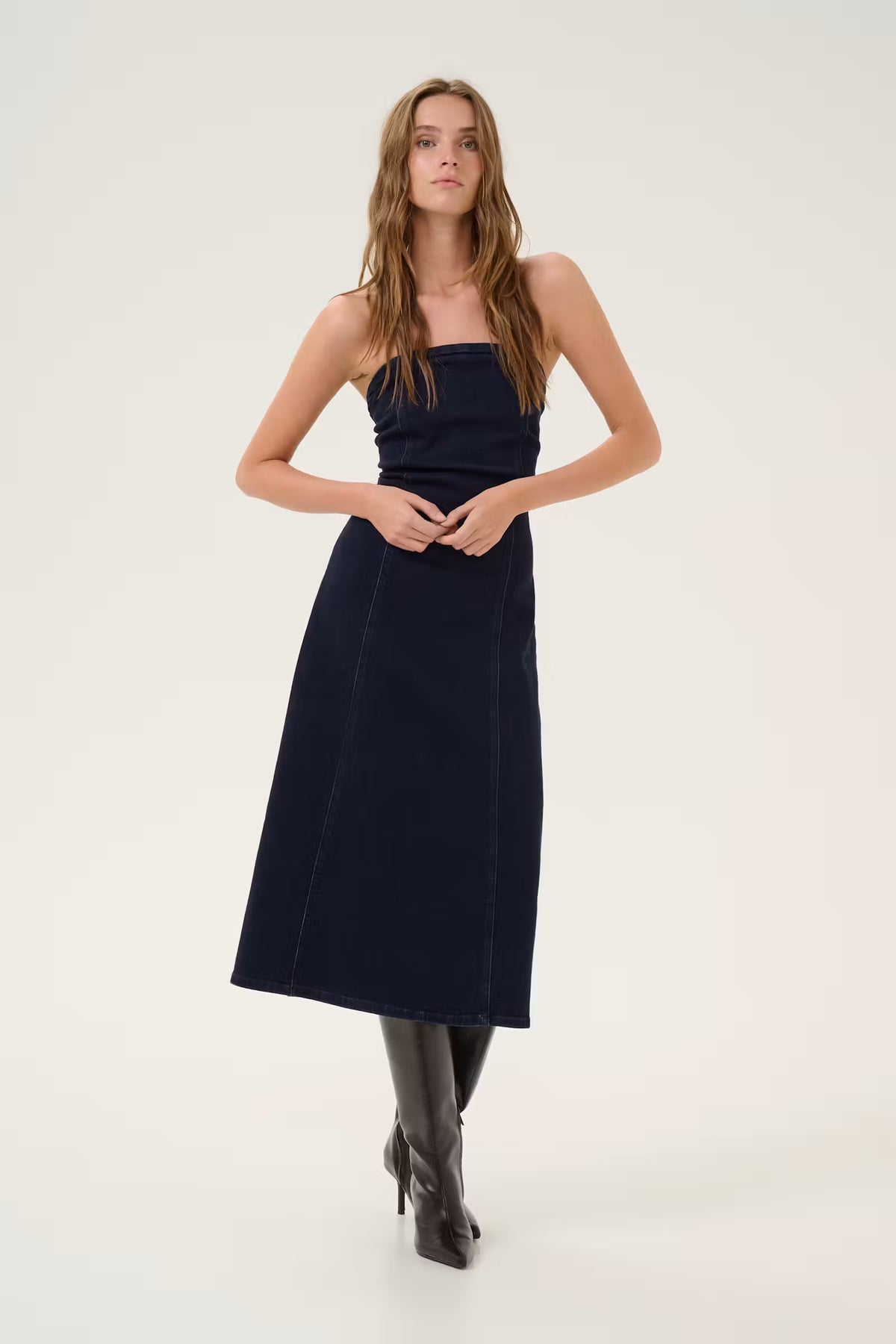 A woman modeling a dark navy blue strapless midi dress with a textured fabric, paired with black knee-high boots, against a plain off-white studio background.