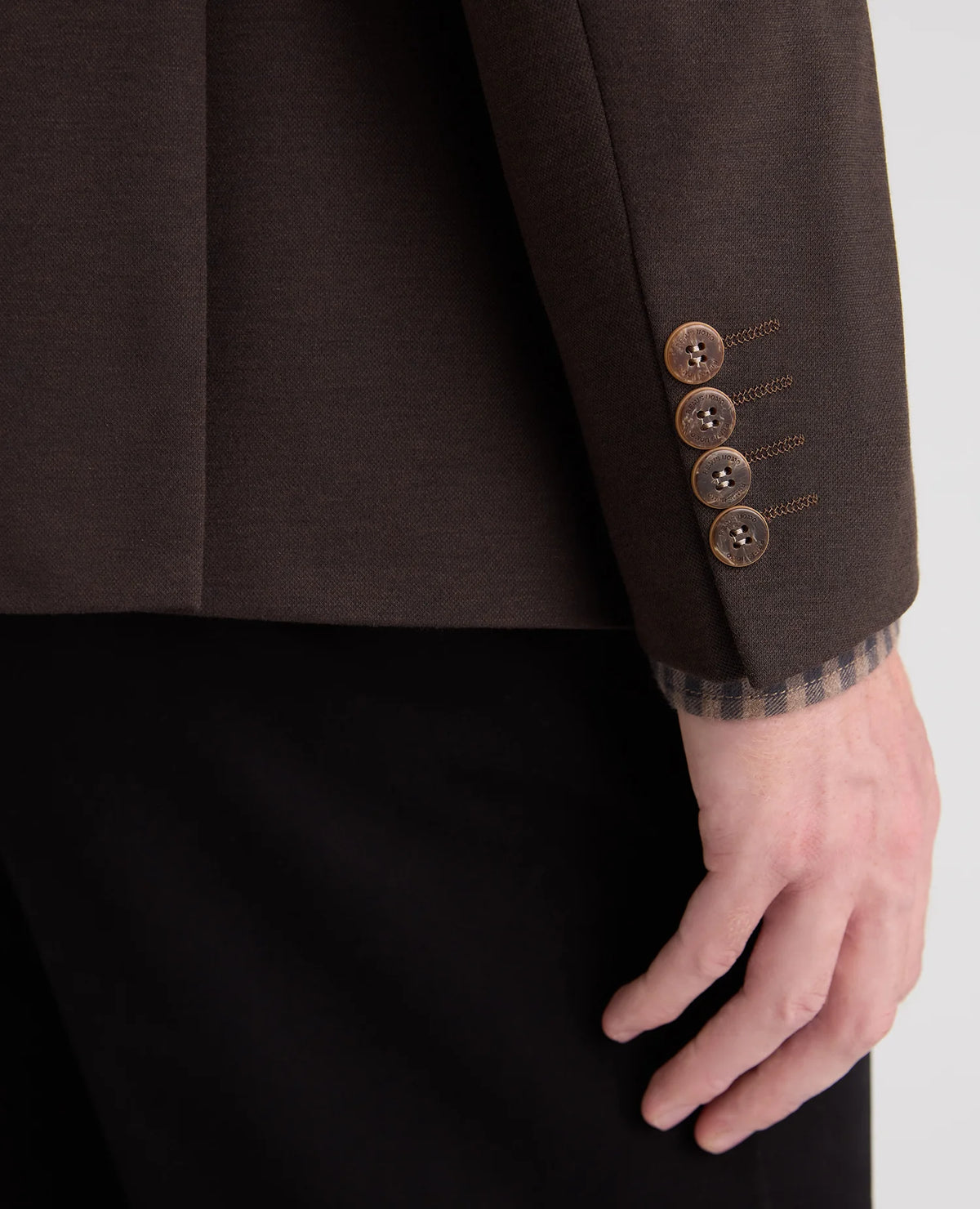 Close-up of a dark brown tailored jacket sleeve with four bronze buttons and a plaid lining at the cuff, worn with black trousers. A hand is visible at the wrist.