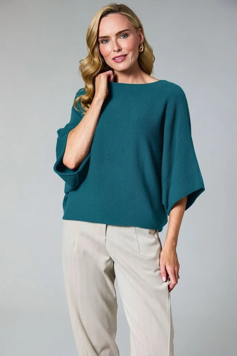 A woman modeling a teal wide-sleeve boatneck top with beige trousers, standing against a neutral gray studio background.