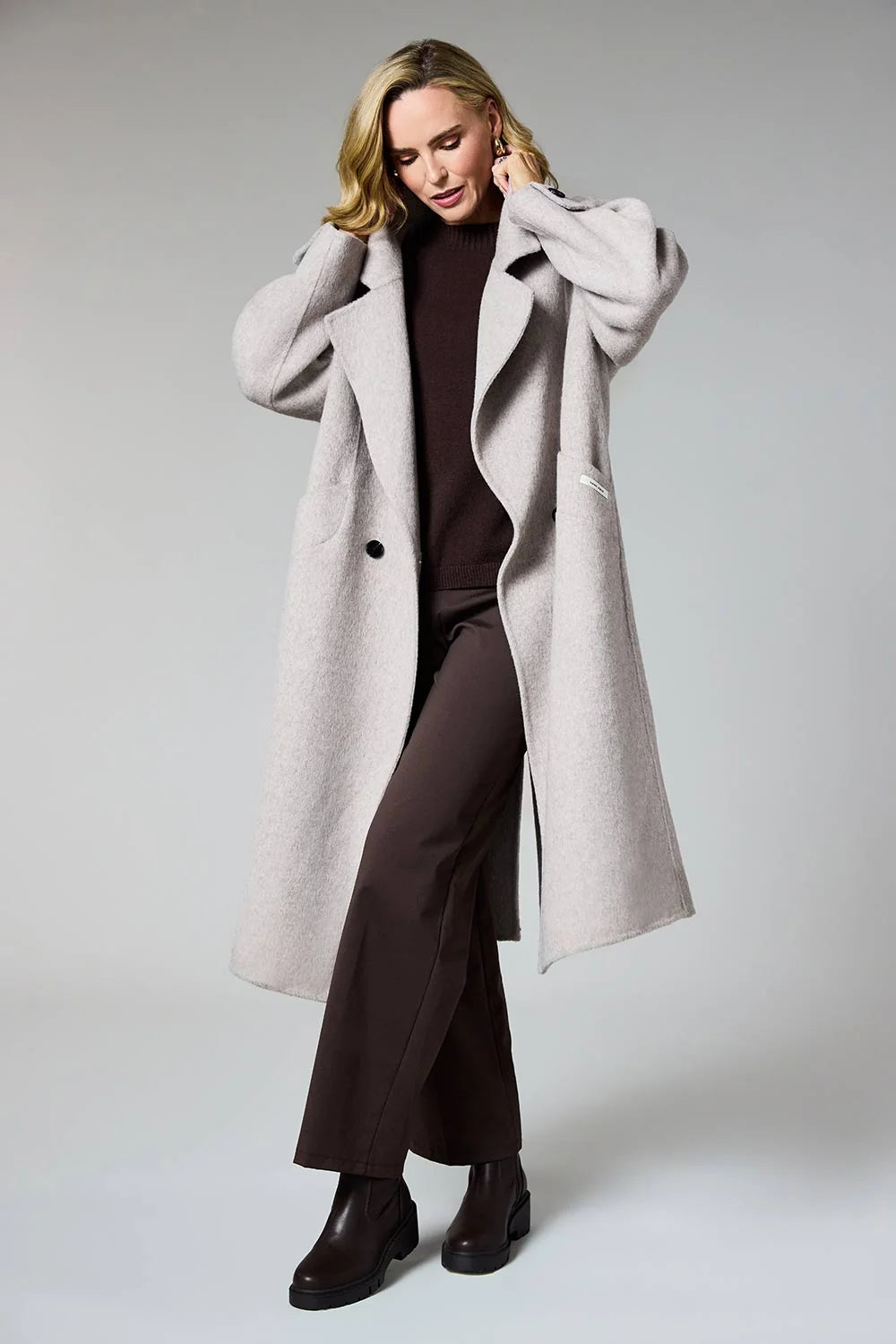A woman modeling a long, light gray wool-blend coat over a dark brown turtleneck and matching wide-leg trousers, standing against a neutral light gray studio background.