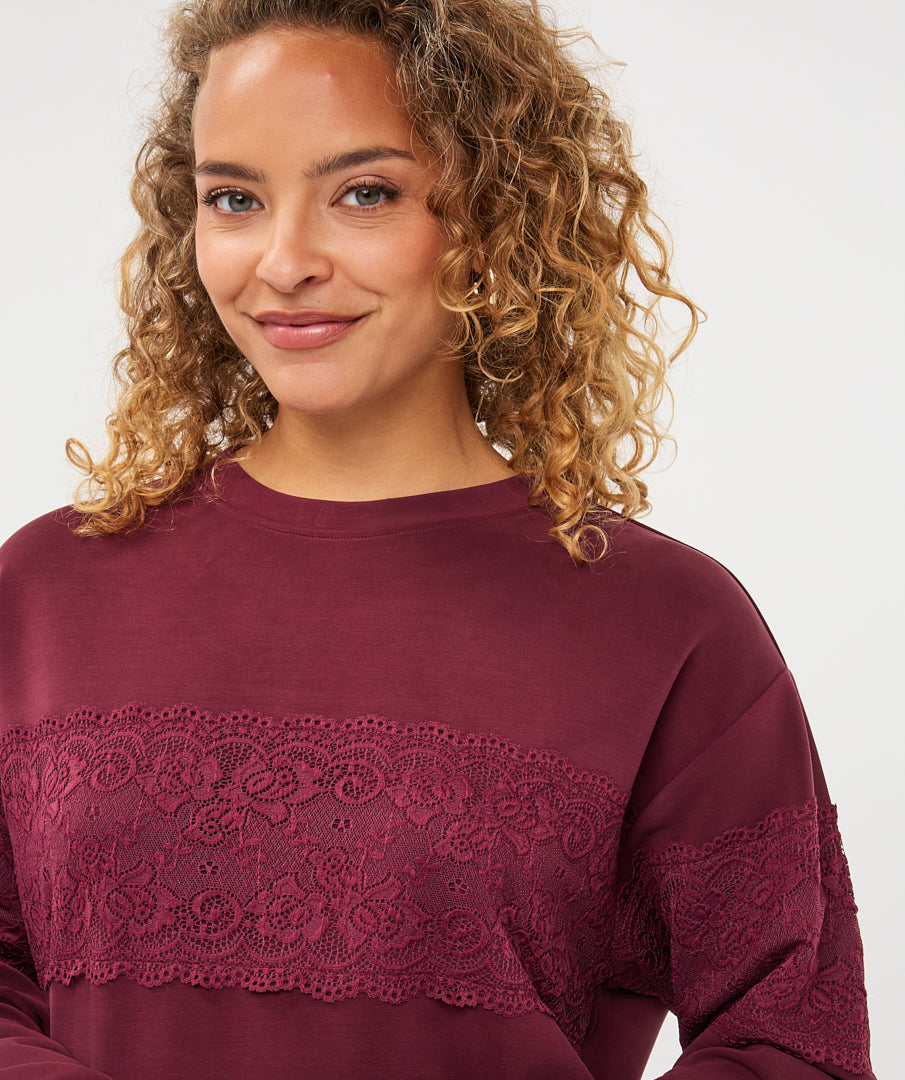 A woman with curly blonde hair wearing a burgundy long-sleeve top with a lace panel across the chest, photographed against a light gray studio background.