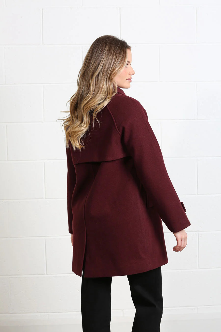 A woman with long blonde hair is shown from the back wearing a mid-length burgundy coat and black pants, standing against a white brick wall.