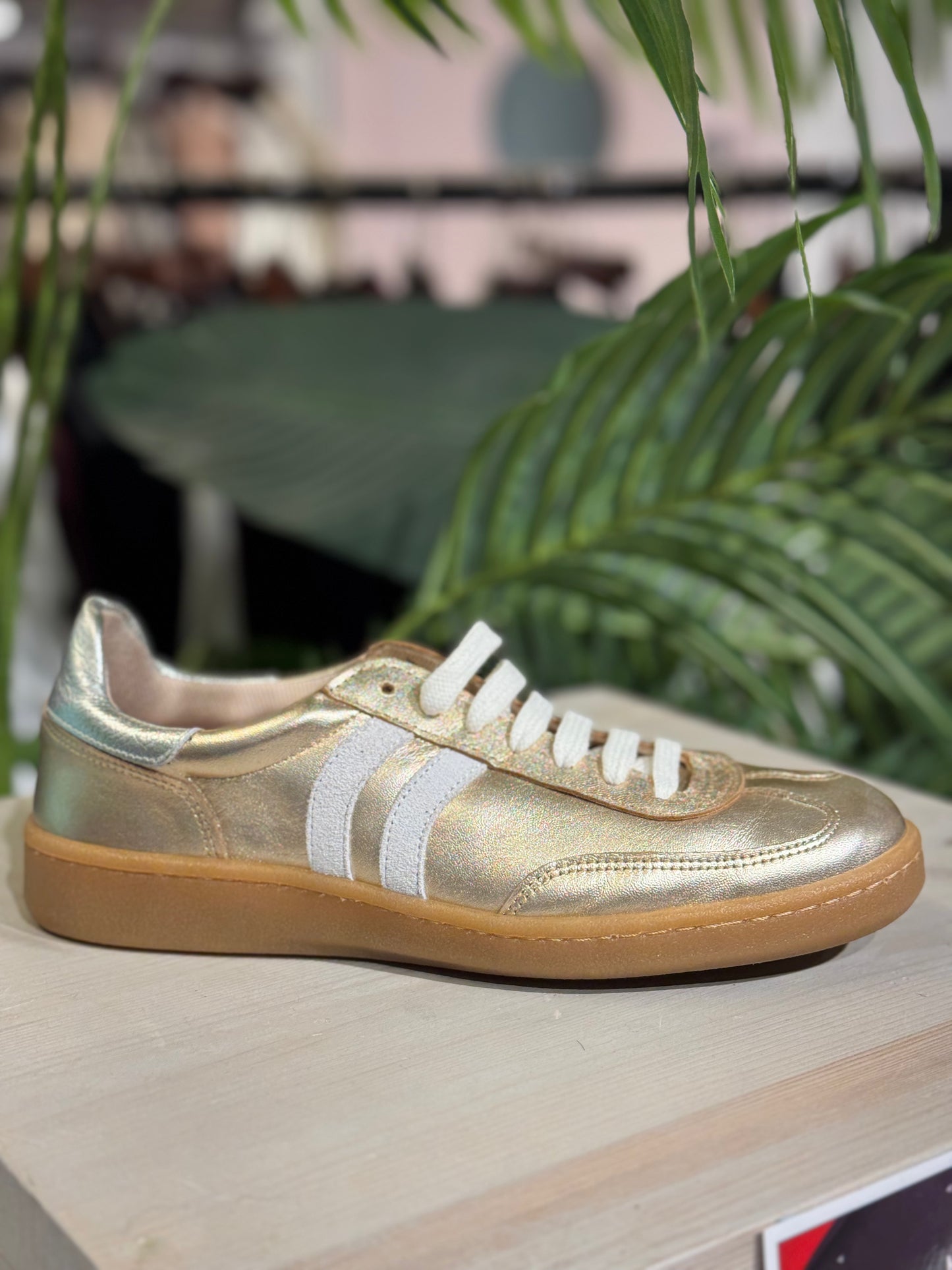Gold sneaker with white laces on a wooden surface with green leaves in the background