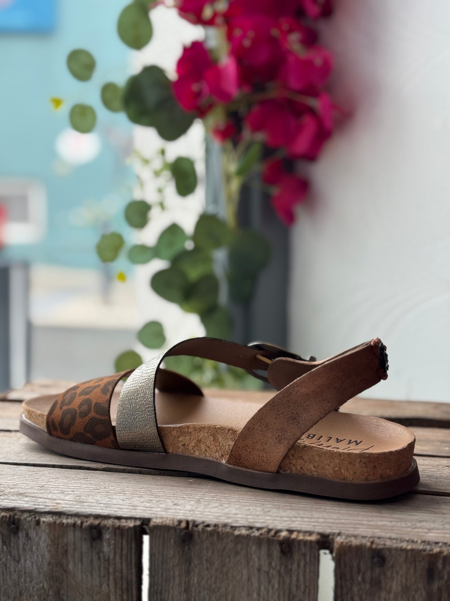 Brown sandal with leopard print strap on a wooden surface with flowers in the background