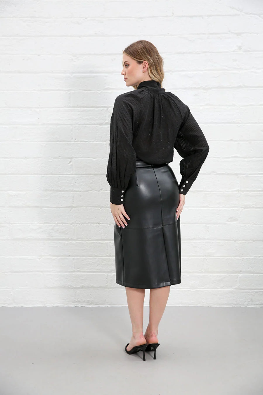 A woman stands with her back to the camera wearing a black long-sleeve blouse and a black leather midi skirt, posed against a white brick wall.