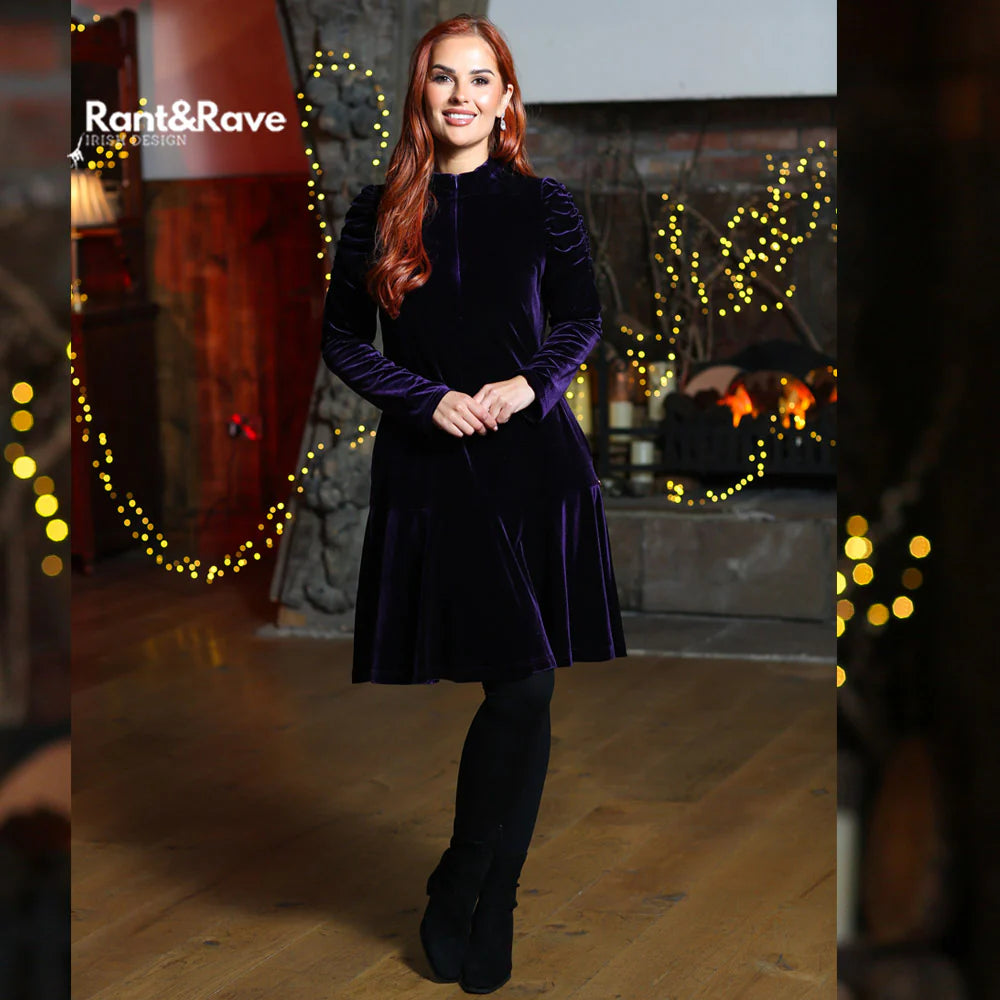 Woman in a purple velvet dress standing in a warmly lit room with decorative lights.