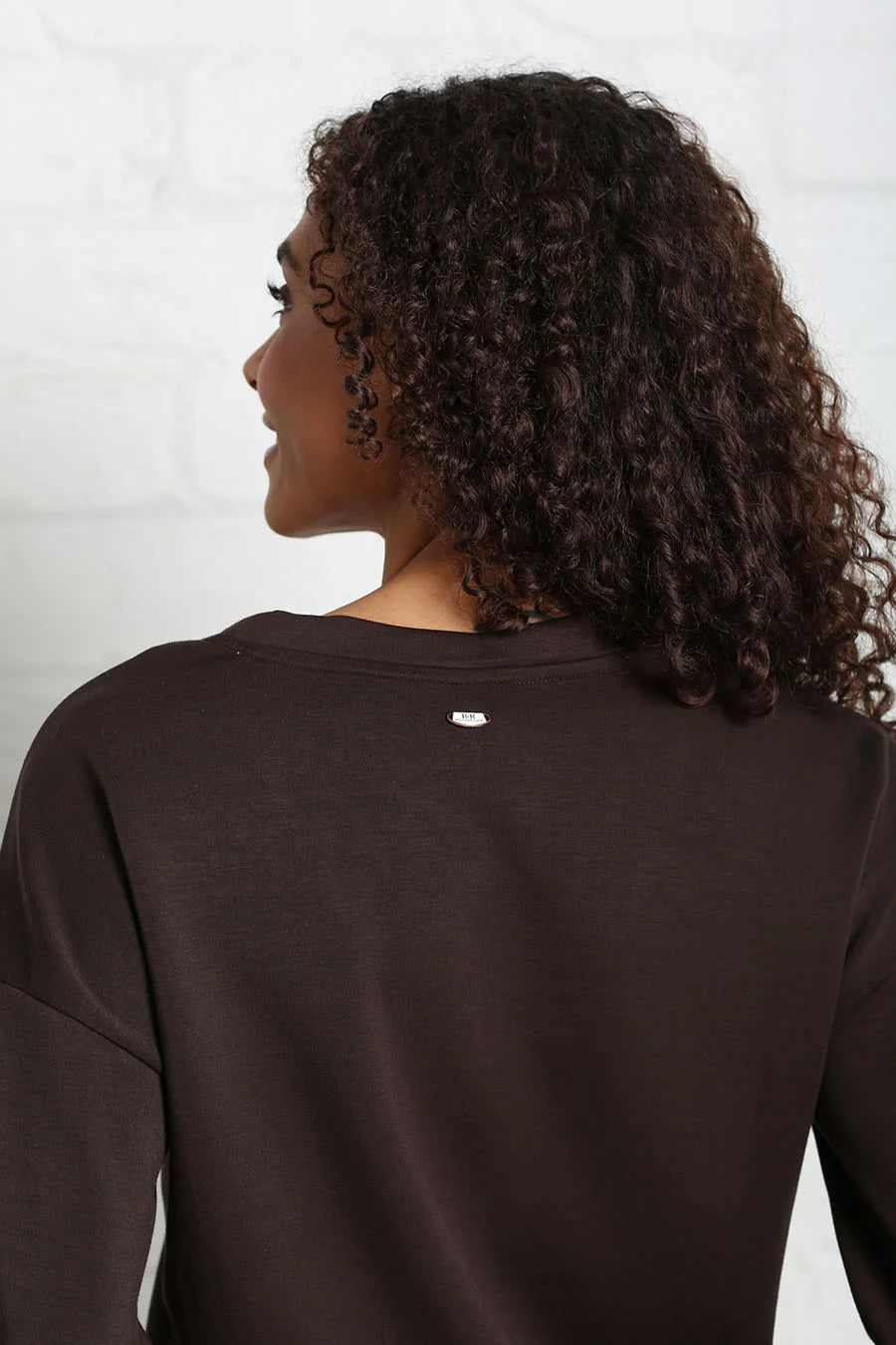 A woman with curly dark brown hair is shown from the back wearing a dark brown crewneck sweatshirt against a white brick wall background.