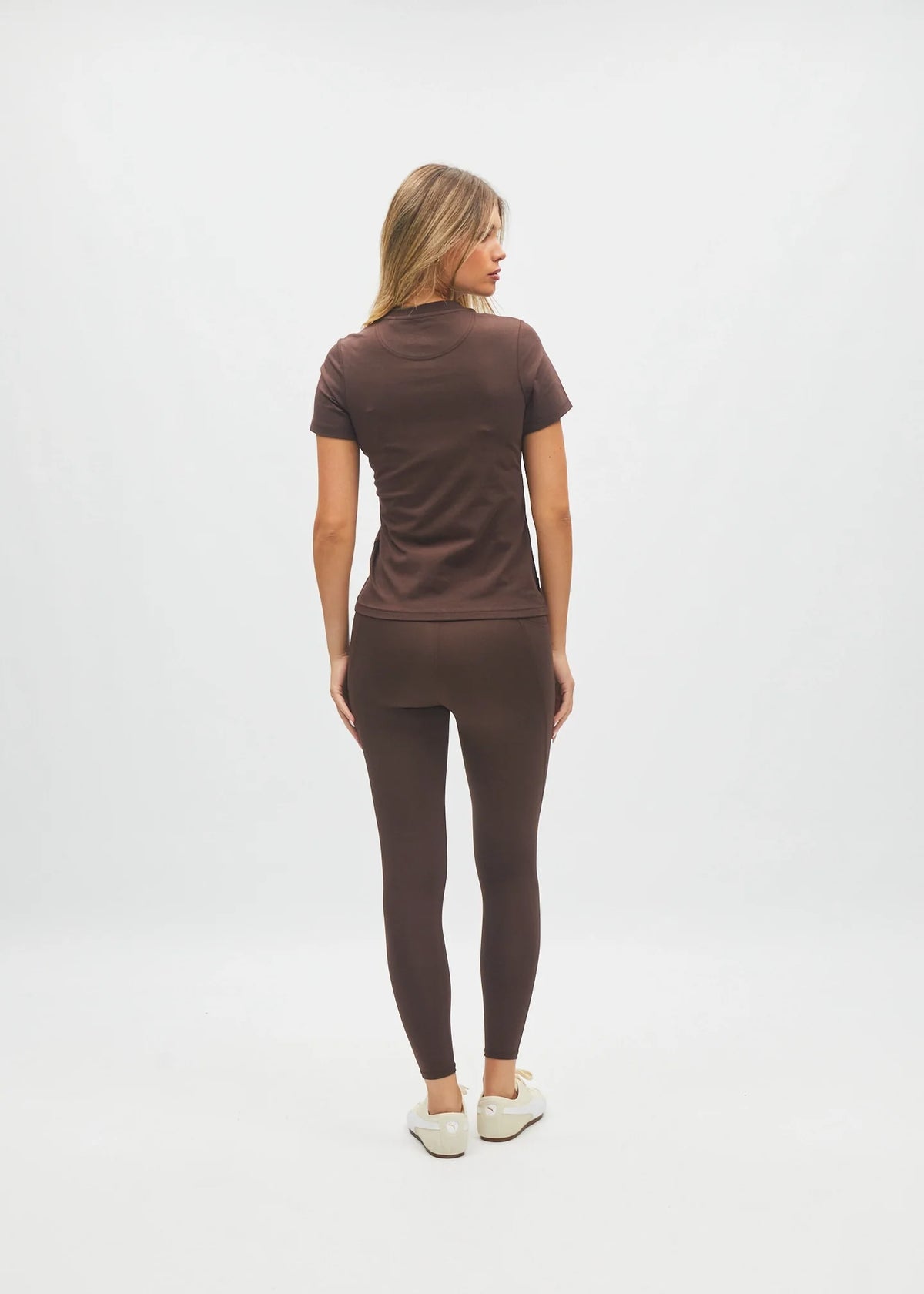 A woman is shown from behind wearing a matching dark brown short-sleeve top and high-waisted leggings set, paired with white sneakers, against a plain white studio background.