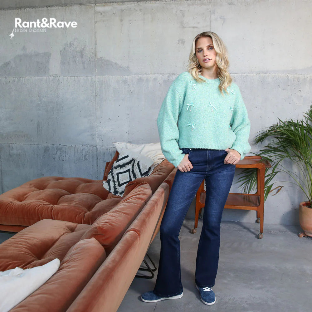 Woman in a light blue sweater and dark jeans standing in a modern living room with a brown sectional sofa.