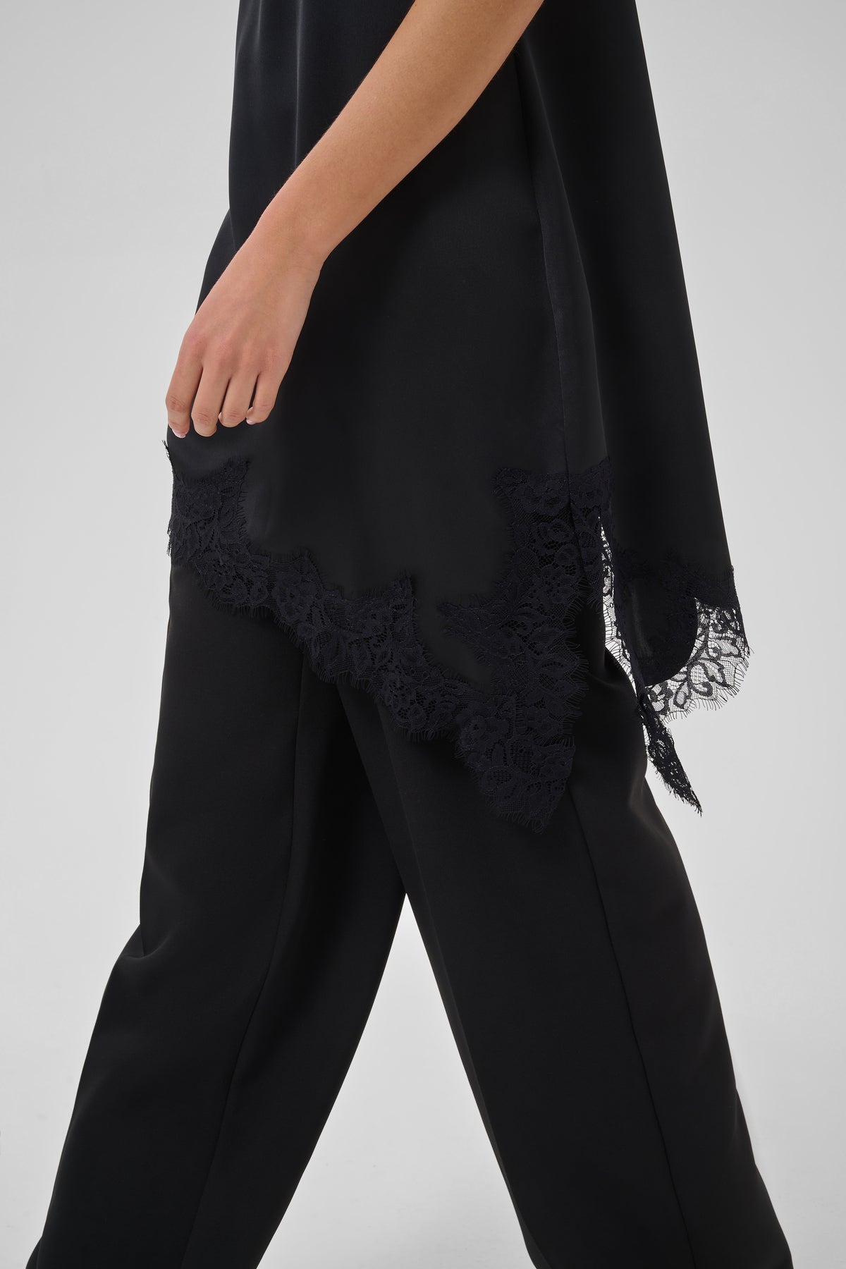 A close-up of a person wearing black wide-leg trousers and a black draped wrap or poncho with lace trim along the hem, photographed against a light gray studio background.