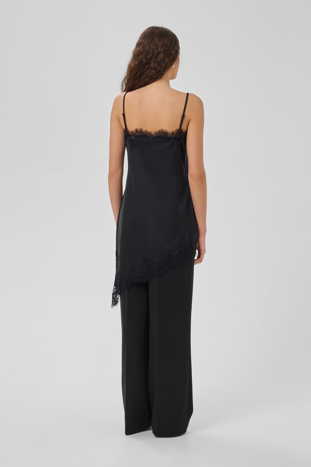 A woman shown from the back wearing a black camisole top with lace trim and wide-leg black pants against a light gray studio background.