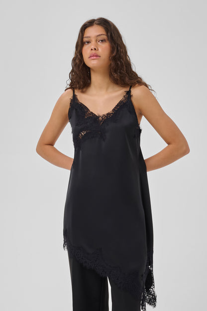 A woman modeling a black satin slip dress with lace trim on the neckline and an asymmetrical lace hem, photographed against a light gray studio background.