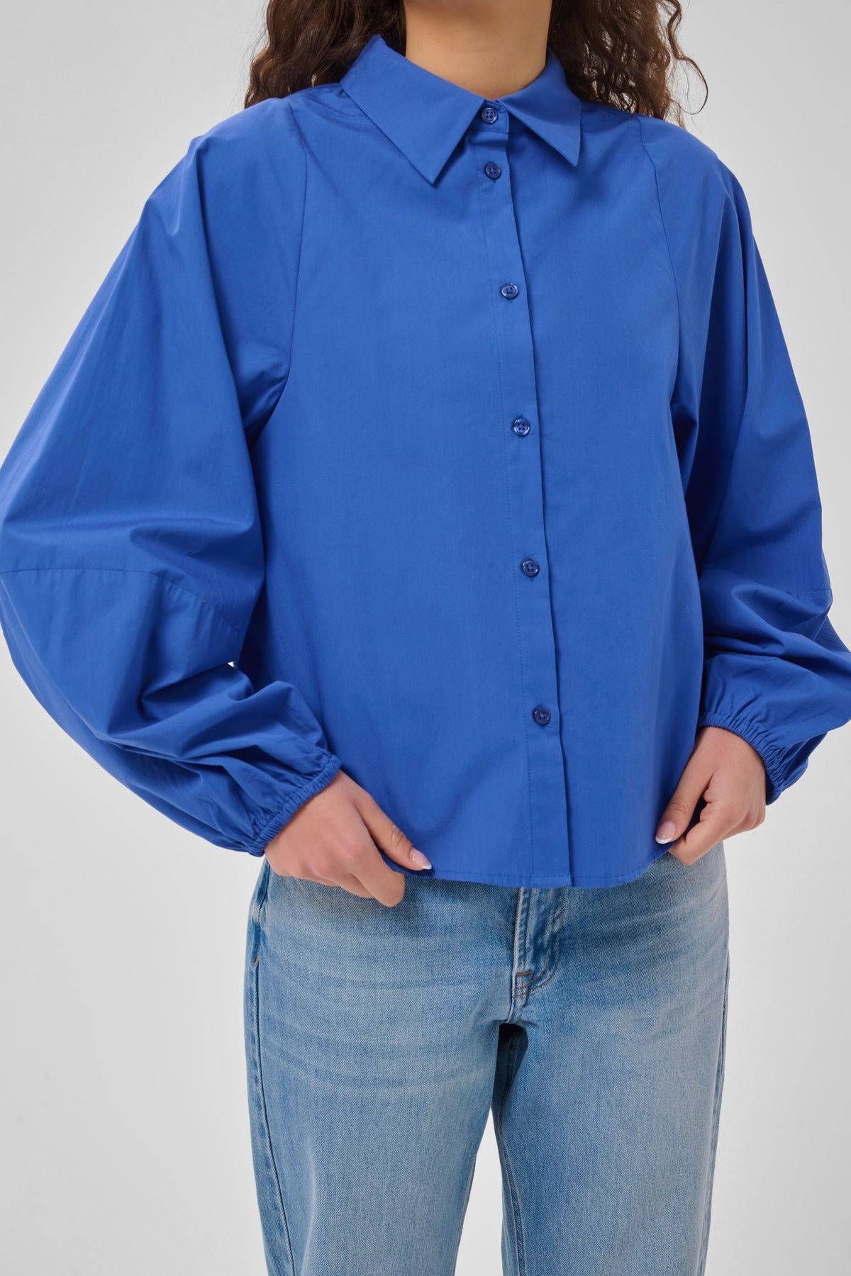 A person wearing a bright royal blue button-front blouse with a pointed collar, wide balloon sleeves, and elasticized cuffs, paired with light blue jeans against a plain light gray studio background.