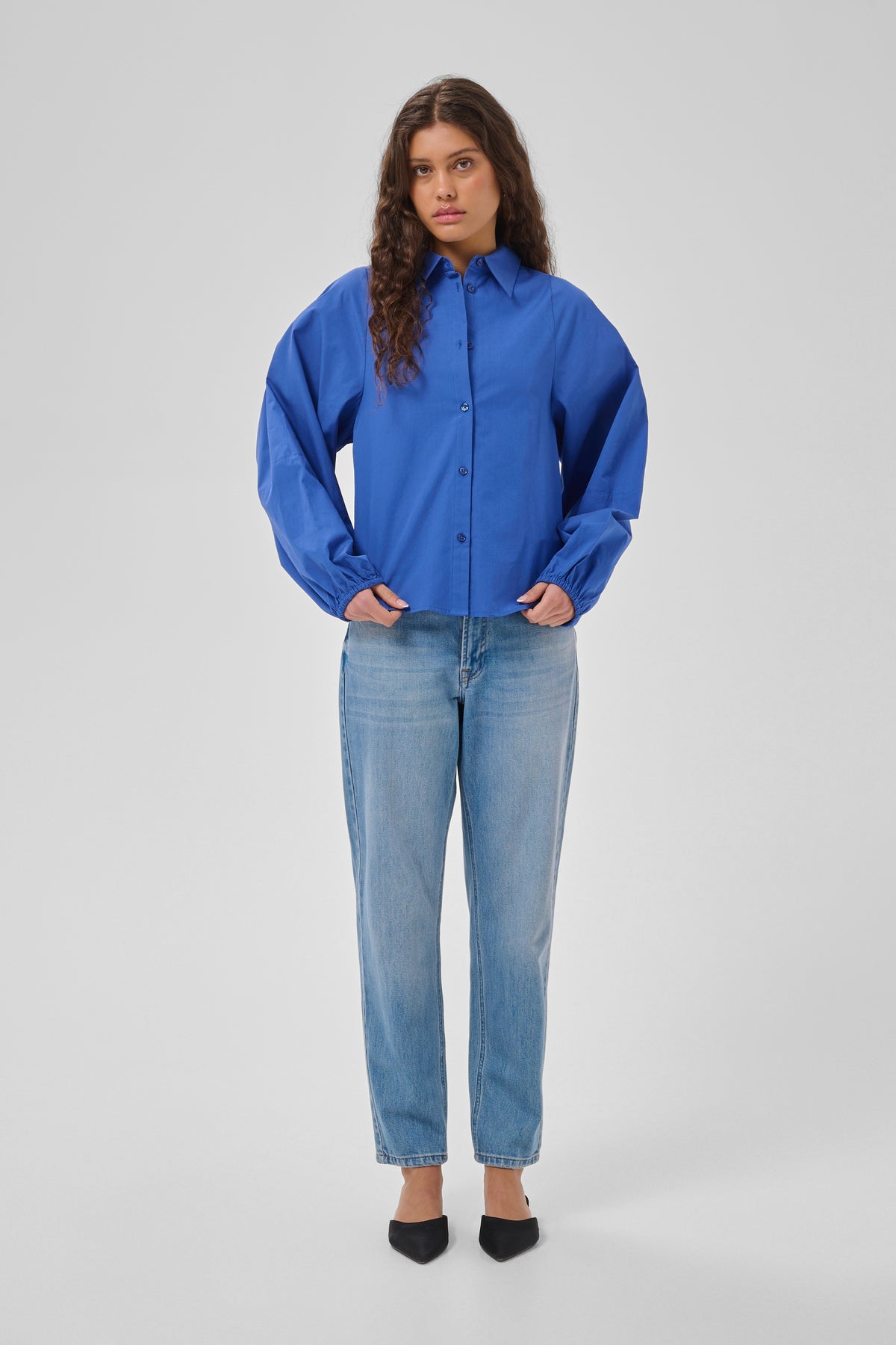 A woman modeling a vibrant royal blue button-up shirt with voluminous balloon sleeves, paired with light-wash blue jeans and black pointed-toe heels, against a neutral light gray studio background.