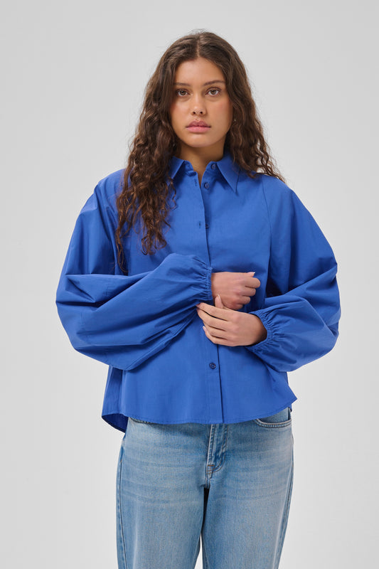 A woman modeling a vibrant royal blue button-up shirt with dramatic, voluminous balloon sleeves and gathered detailing, paired with light-wash blue jeans against a neutral gray studio background.