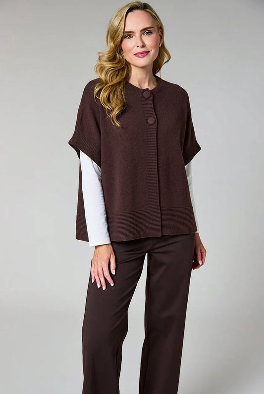 A woman modeling a dark brown knit poncho-style top with a button front and white long-sleeve undersleeves, paired with matching dark brown pants, against a light gray studio background.