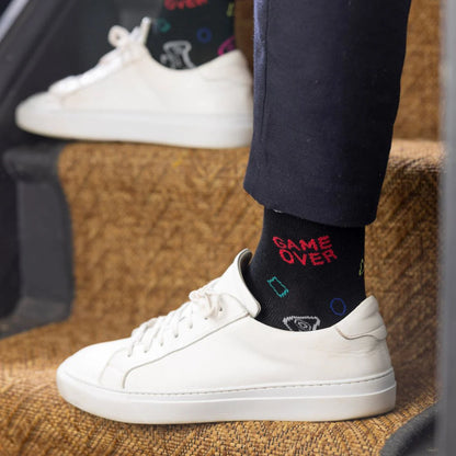 Person wearing black socks with 'GAME OVER' text and white sneakers on a textured surface.