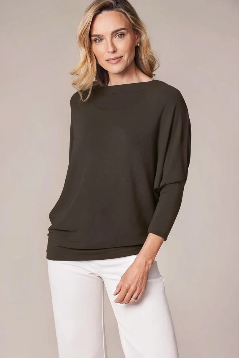A woman modeling a dark brown boat-neck knit top with elbow-length sleeves and white pants, photographed against a plain beige studio background.