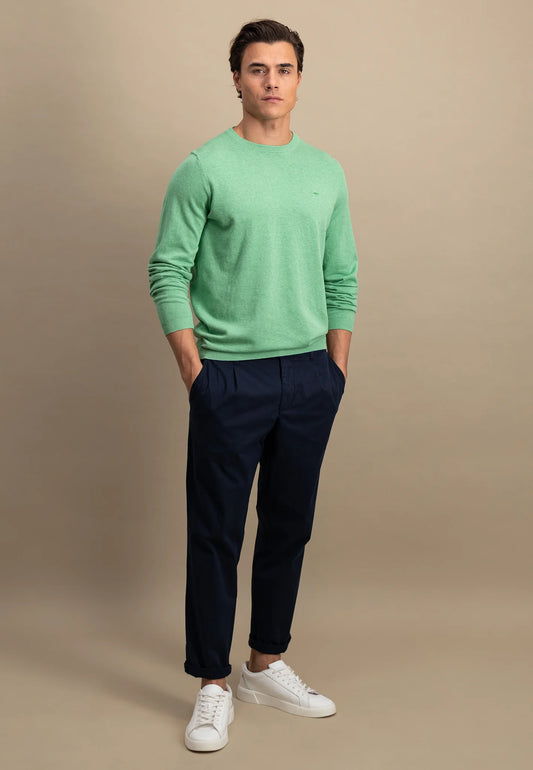 A man wearing a turf green round neck jumper and navy pants with white shoes standing against a beige background.