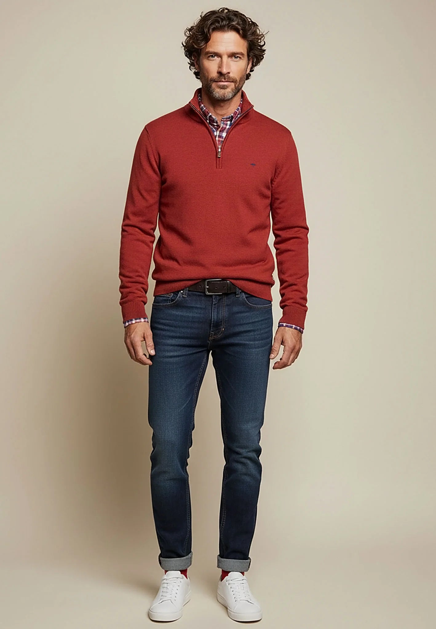 Man wearing a red sweater, blue jeans, and white sneakers against a beige background
