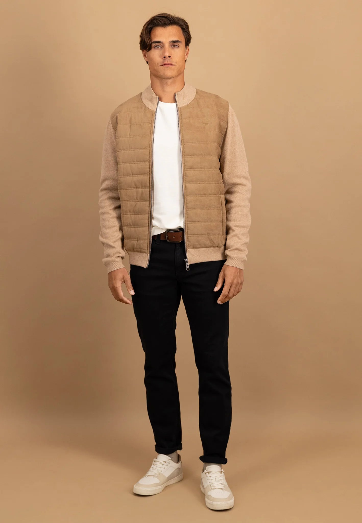 Man wearing a beige quilted jacket, white shirt, black pants, and white sneakers on a brown background