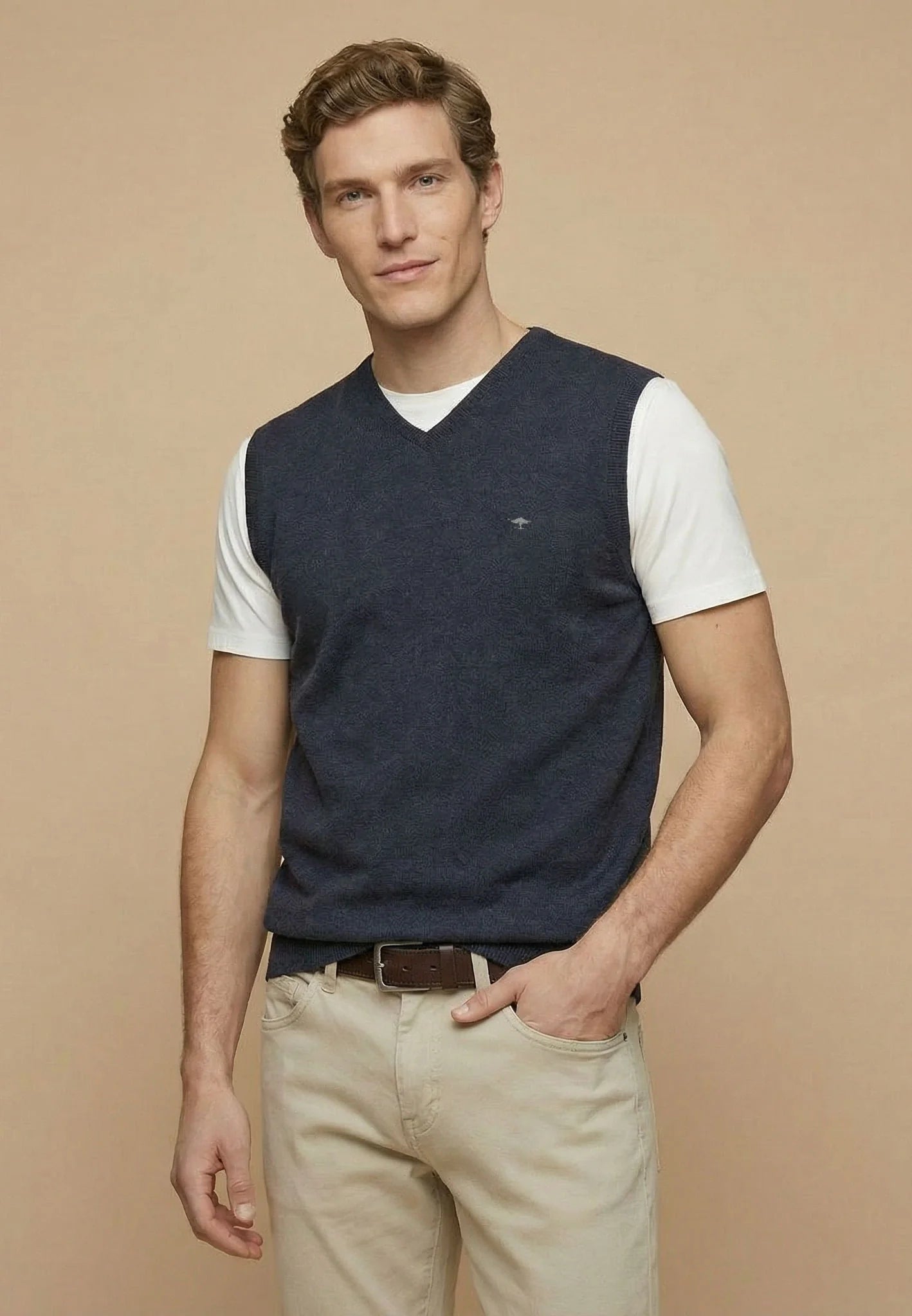 Man wearing a navy sweater vest over a white shirt with beige pants on a beige background