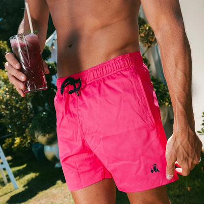 Person wearing bright pink swim shorts holding a drink outdoors.