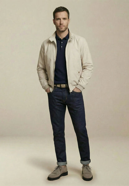Man wearing a beige jacket, navy shirt, and dark jeans on a plain background