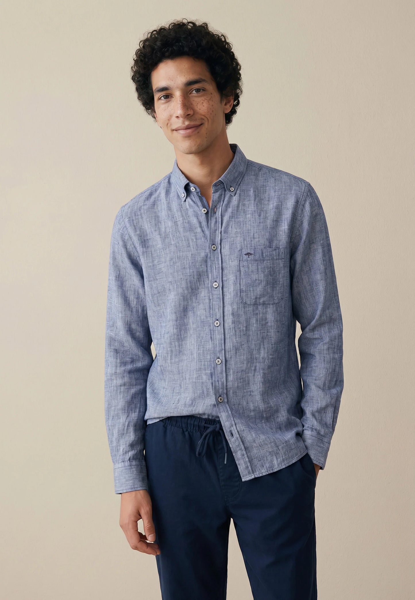 Man wearing a blue button-up shirt and dark pants against a beige background