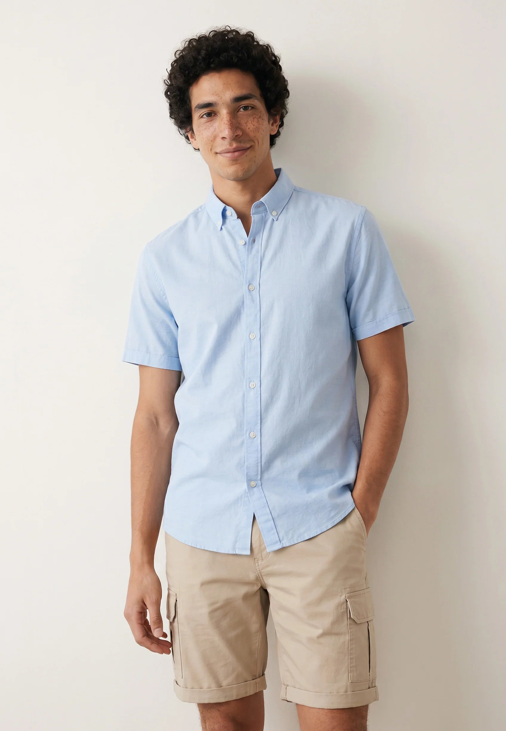 Man wearing a light blue shirt and beige shorts against a plain background