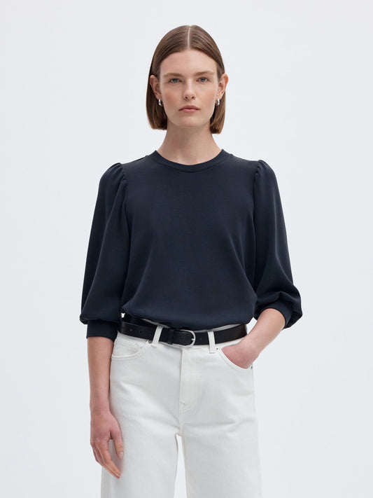 A model wearing a dark navy blue long-sleeve blouse with puffed shoulders, tucked into white trousers with a black belt, against a light gray studio background.