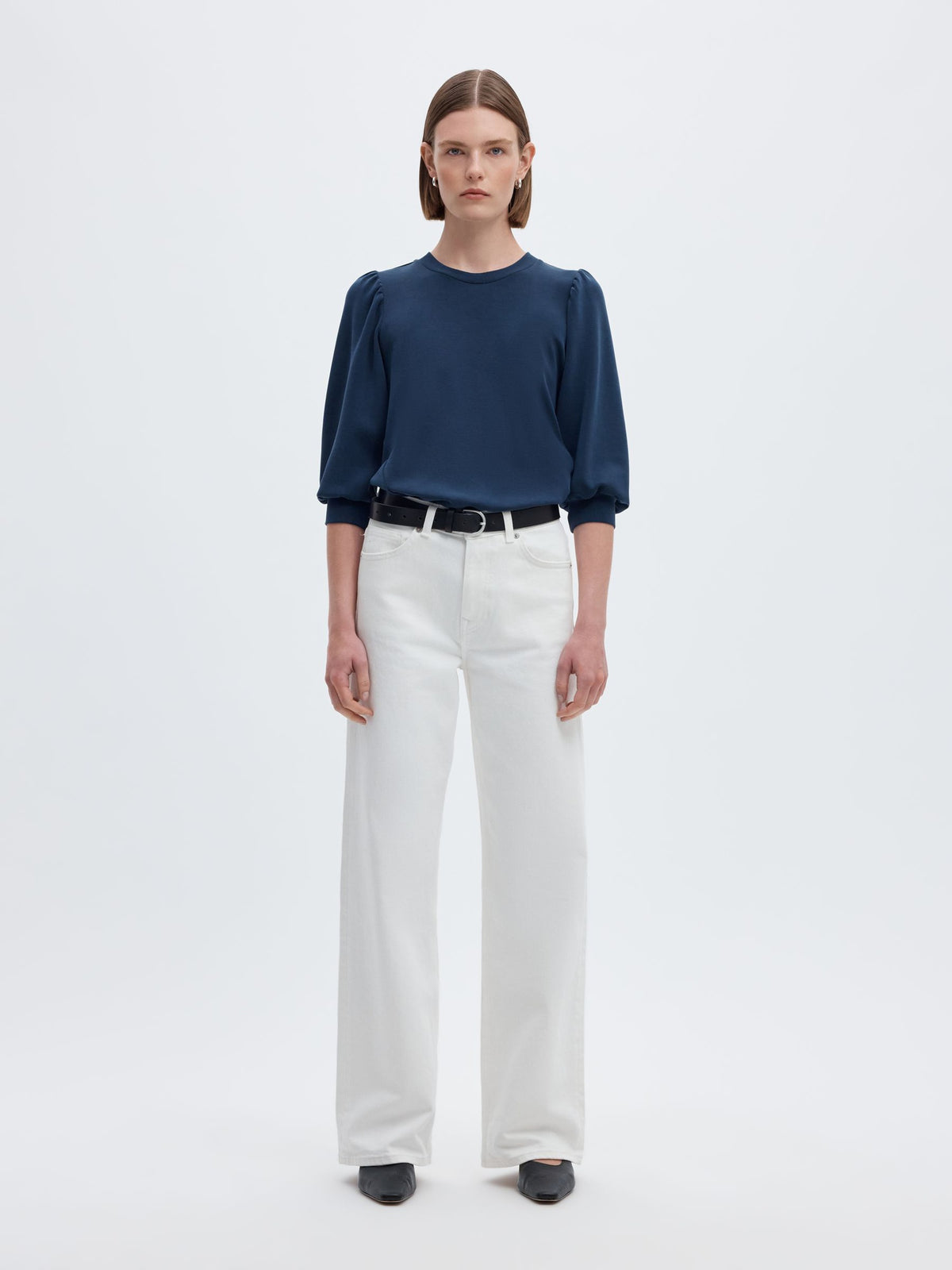 A model stands against a light gray studio background wearing a navy blue long-sleeve blouse with puffed shoulders, white wide-leg trousers, a black belt, and black shoes.