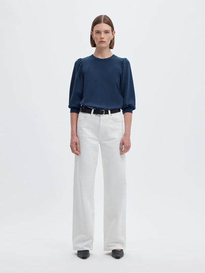 A model stands against a light gray studio background wearing a navy blue long-sleeve blouse with puffed shoulders, white wide-leg trousers, a black belt, and black shoes.