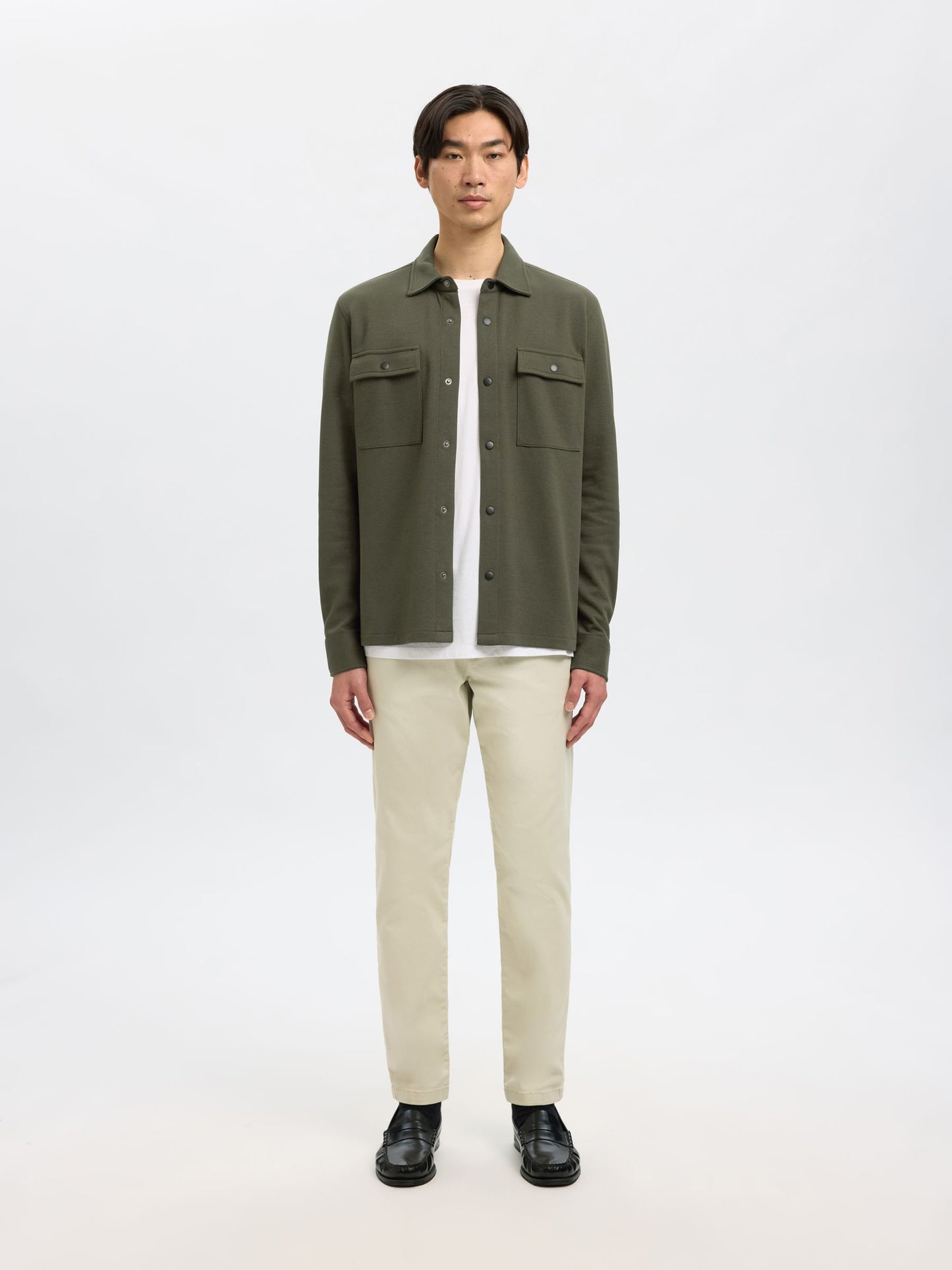 A man modeling an olive green button-up shirt jacket over a white t-shirt, paired with beige trousers and black shoes, against a plain white studio background.