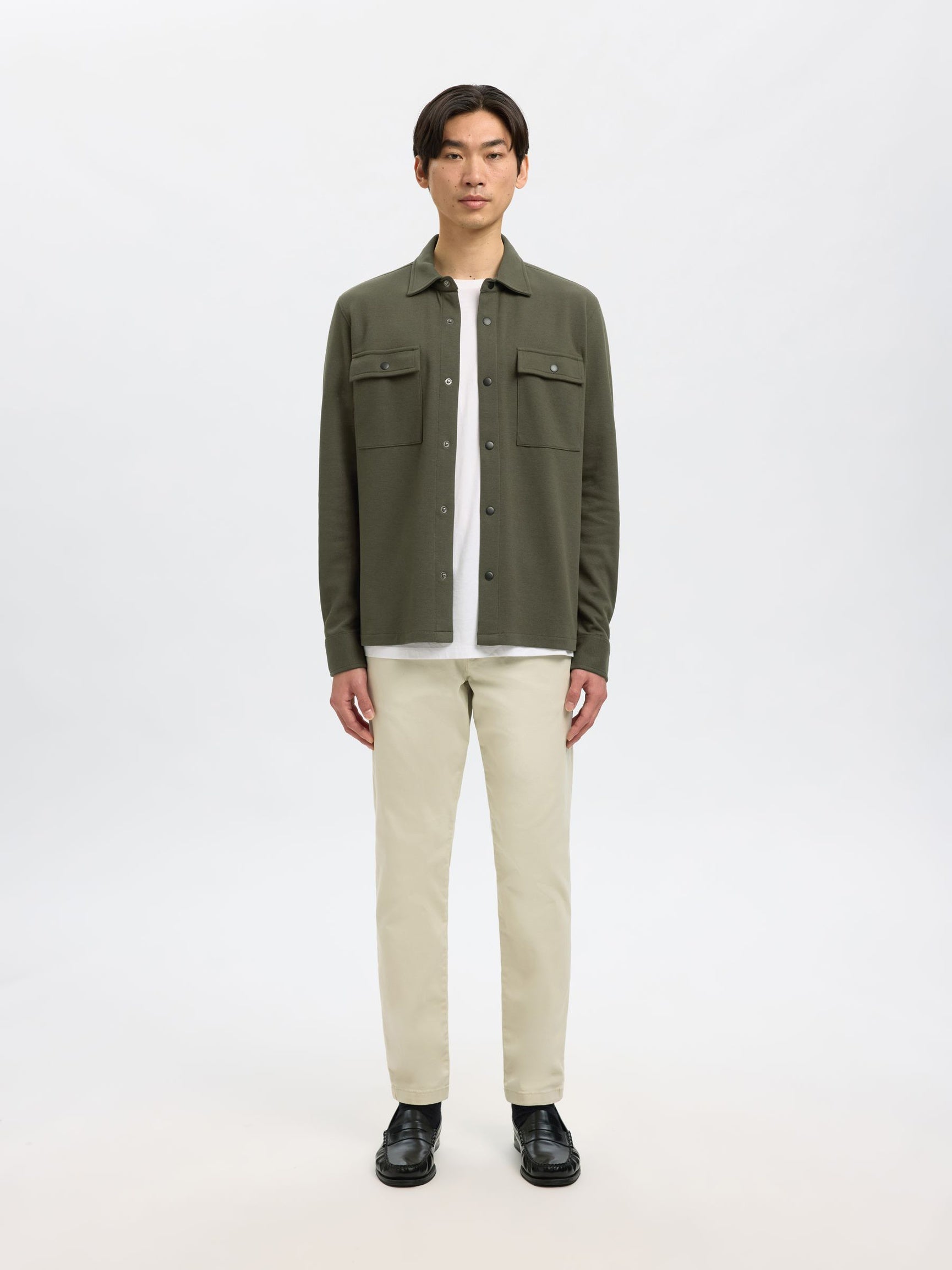 A man modeling an olive green button-up shirt jacket over a white t-shirt, paired with beige trousers and black shoes, against a plain white studio background.