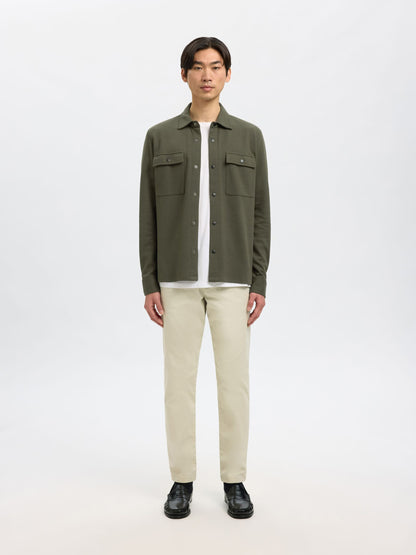 A man modeling an olive green button-up shirt jacket over a white t-shirt, paired with beige trousers and black shoes, against a plain white studio background.