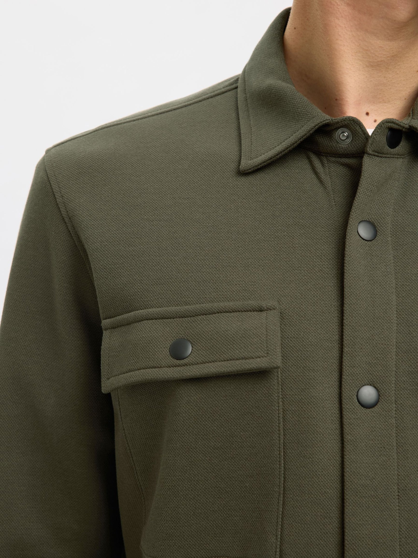 Close-up of a person wearing an olive green textured overshirt with a button-down front, pointed collar, and a chest pocket with a snap tab.