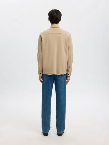 A man viewed from behind wearing a beige long-sleeve button-up shirt and blue jeans, standing against a plain white studio background.