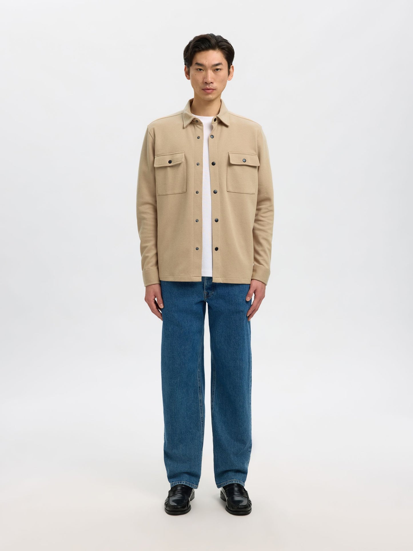 A man stands against a light gray studio background wearing a beige button-up overshirt over a white T-shirt, blue jeans, and black shoes.